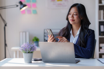 Young office worker is sitting at her desk, using a smartphone and working with her laptop, focusing on her tasks in a bright and modern workspace