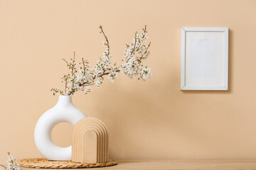 Vase with blossoming branches and blank frame on beige wall