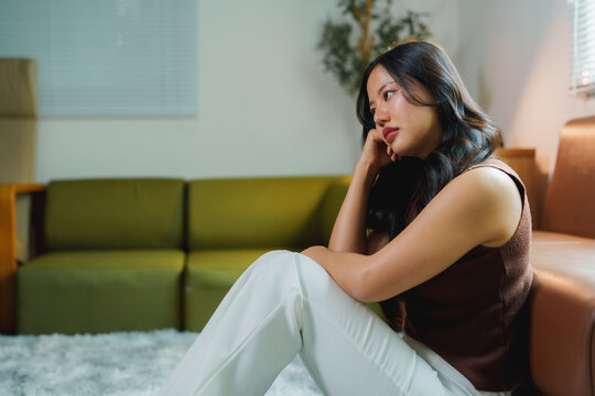 Young asian woman sitting on floor near sofa at home looking away with thoughtful and sad expression, suffering from psychological problems, feeling lonely and depressed