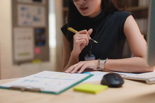 Businesswoman wearing a smartwatch and holding a pen, analyzing a financial report while seated at her desk in a sleek, modern office environment, focused on data-driven decision making