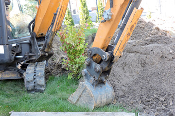 A narrow bucket, a dipper and hydraulic cylinders of a black and yellow mini hydraulic excavator standing in a courtyard next to heap of dirt