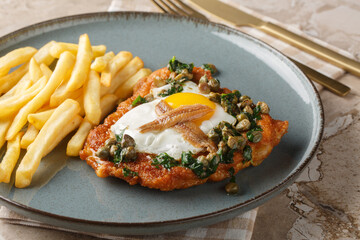 Delicious breaded schnitzel with anchovies, fried egg and capers parsley sauce on top served with french fries close-up on a plate on the table. Horizontal