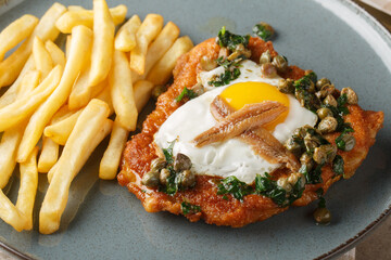 Schnitzel Holstein consists of a breaded cutlet served with a fried egg, anchovy and lemony caper sauce on top close up on the plate on the table. Horizontal