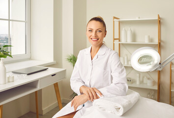 Portrait of a happy young smiling woman cosmetologist or beautician doctor sitting in a beauty spa salon office on her workplace and looking cheerful at camera. Cosmetology and skincare concept.