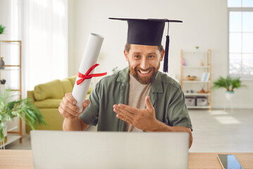 Happy cheerful smiling man completes online business elearning degree, sits in student cap at desk,...