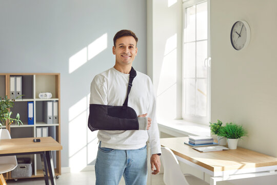 Portrait of a young smiling man standing at home or at office on rehabilitation with his arm in a plaster cast after injury. Patient wearing sling or bandage. Recovery and rehab concept.