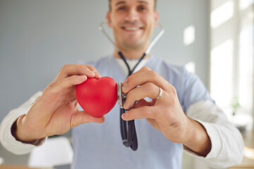 Close up hands of male smiling young doctor cardiologist with stethoscope examining red heart...