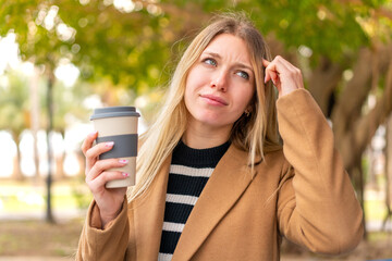 Young pretty blonde woman holding a take away coffee at outdoors having doubts and with confuse...