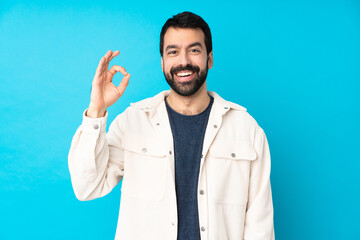 Fototapeta premium Young handsome man with white corduroy jacket over isolated blue background surprised and showing ok sign