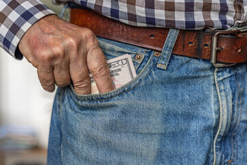 Tight close-up of a man's fingers inserting or pulling a wad of 50 American dollar banknotes from his jeans front pocket. Cash in hand concept - wages, payment, corruption, dividends or loan.