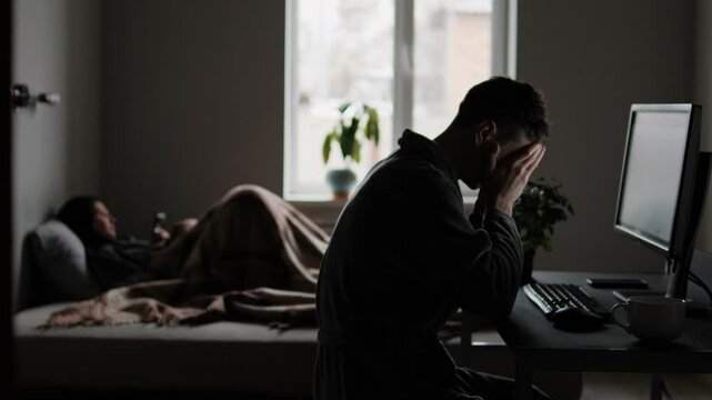 Man sitting at desk, wearing robe, holding head in despair in front of monitor after financial loss, while woman lies on bed in background scrolling phone, distant and indifferent. Dark home interior