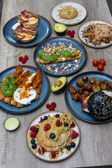 Colorful modern brunch table with avocado toast, eggs, fruit pancakes, black burger and plantains flat lay
