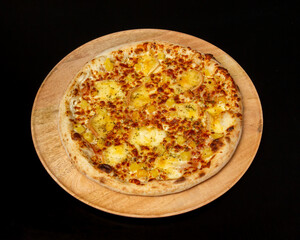 Overhead view of a pineapple pizza on a wooden plate against black background