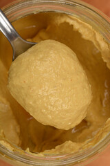 A spoonful of creamy yellow mustard is held above a partially filled glass jar of the same condiment