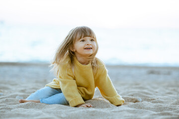 Smiling kid girl 2-3 year old wearing yellow knitted sweater at sandy beach over sea background. Summer season. Childhood.