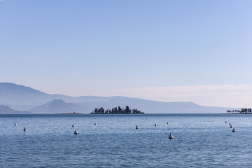 Obraz premium Peaceful spring morning on Lake Garda with distant San Biagio island and blue mountain silhouettes fading into gentle haze over calm water