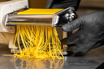 A chef's gloved hand guides fresh yellow pasta through a pasta maker, forming long, delicate strands. The stainless steel surface reflects the artistry of traditional pasta preparation.