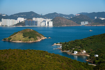 Panoramic view of the Hong Kong coastline with green forest in the foreground and high-rise residential buildings nestled at the base of lush hills under a bright blue sky