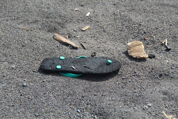 A worn-out black flip-flop with teal straps lies abandoned on a sandy surface with scattered...