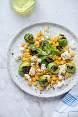 Plate with sweet corn, grilled Brussels sprouts and cheese, vertical shot on a white stone background, elevated view