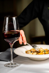 A hand gracefully uses a fork to savor fettuccine pasta with chicken and mushrooms, accompanied by a glass of red wine. The elegant setting enhances the allure of the delicious gourmet meal.
