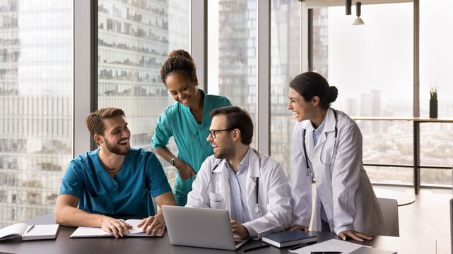 Joyful collaboration. Happy multiethnic doctors team gather around notebook at modern meeting room make funny comments reviewing notes presentations online take break in work to watch hilarious video