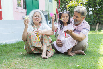 Indian grandparents with granddaughter blow soap bubbles outdoor. Family with girl child. Summer activity.