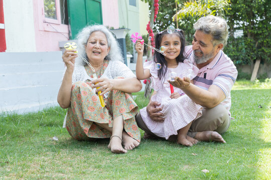 Indian grandparents with granddaughter blow soap bubbles outdoor. Family with girl child. Summer activity.