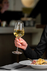 A hand holds a glass of white wine, capturing the light beautifully. In the background, a elegantly set table and a plate of spaghetti suggest a refined dining atmosphere.