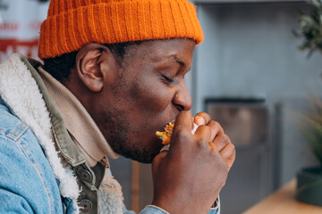 Young african american man wearing bright orange winter beanie, biting into juicy burger inside casual fast food restaurant, side profile capturing winter street food moment