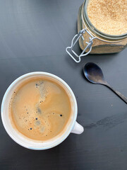 Close-up of a cup of fresh coffee with a frothy crema, alongside a jar of sugar and a spoon.