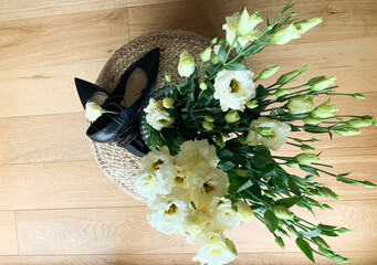 Top view of a bouquet with white lisianthus flowers in a vase next to elegant black shoes on a woven stool, placed on wooden floor.
