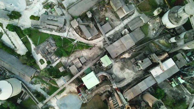 Aerial view of an old cement factory surrounded by greenery. The industrial site features tall silos, factory buildings, and nearby railway tracks.