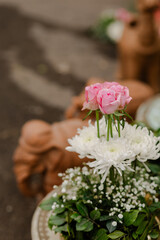 Floral arrangement of pink roses, white daisies, and baby's breath in a blurred outdoor setting. A terracotta figure adds a rustic touch.