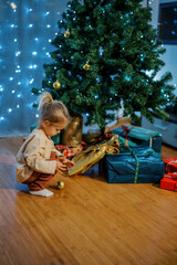 Little girl squat down near the Christmas tree to pick up a golden ball from the floor