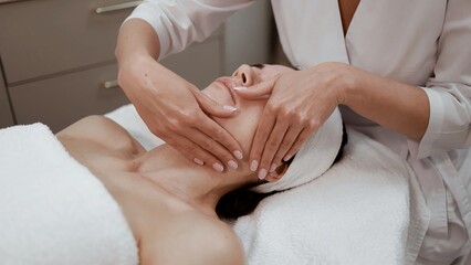 Young woman lying on bed at spa salon and enjoying pampering facial massage. Cosmetologist...
