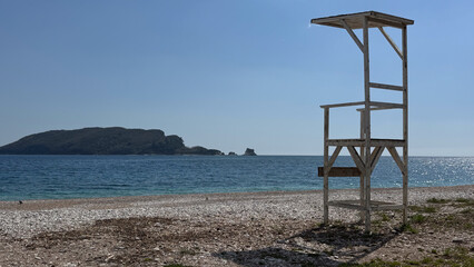 A lone wooden lifeguard tower stands on a deserted pebble beach with a calm sea and blue sky in the background.