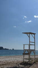 A lone wooden lifeguard tower stands on a deserted pebble beach with a calm sea and blue sky in the background.