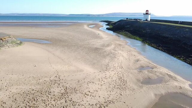 Drone shot travelling forwards from the entrance to Bury Port Harbour, along the breakwater, over the beach and past the lighthouse. Burry Port is in Caramarthenshire in South Wales, UK. A sunny sprin