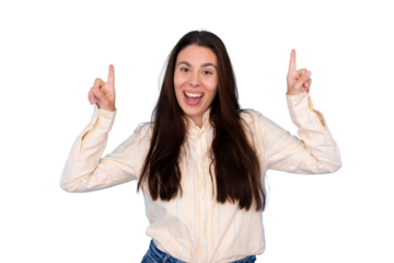 Young woman with long brown hair wearing a beige shirt is pointing up with both index fingers and smiling excitedly on a transparent background