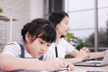 Smiling Asian mother working with laptop and daughter writing with pencil on book together in office room at home. Child educational for homeschooling concept