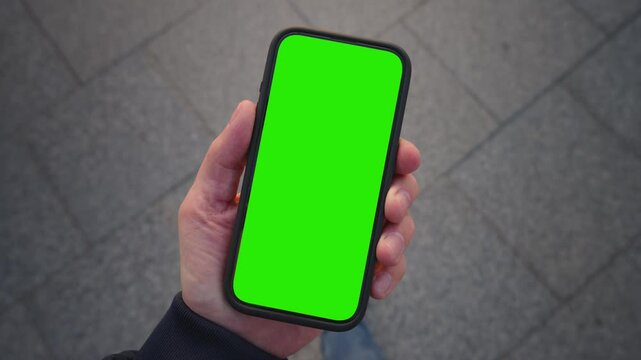 POV shot caucasian man looking at chroma key phone in his hand walking city street. Smart phone green screen in hand. Walk with cellphone, application mock up, camera locked, paving slabs background