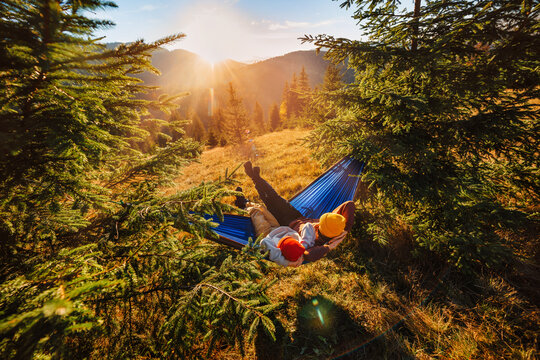 Couple of tourists resting in a nylon hammock hanging between fir trees, mountain landscape background at sunset. Family camping in the mountains. Wide angle shooting.