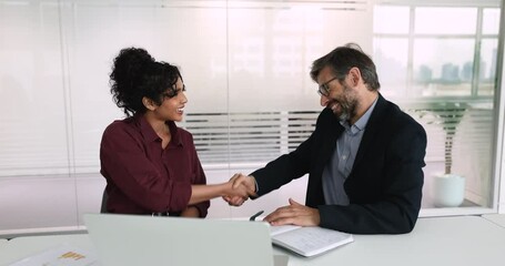 Smiling Indian businesswoman shaking hands with businessman seated at meeting table, closing commercial deal, start partnership, concluding successful job interview or negotiation. Business, etiquette