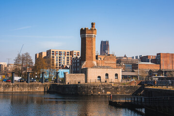 Fototapeta premium Waterfront scene in Liverpool featuring a historic brick tower, modern and historic buildings, the Liverpool Cathedral, and calm reflective water.
