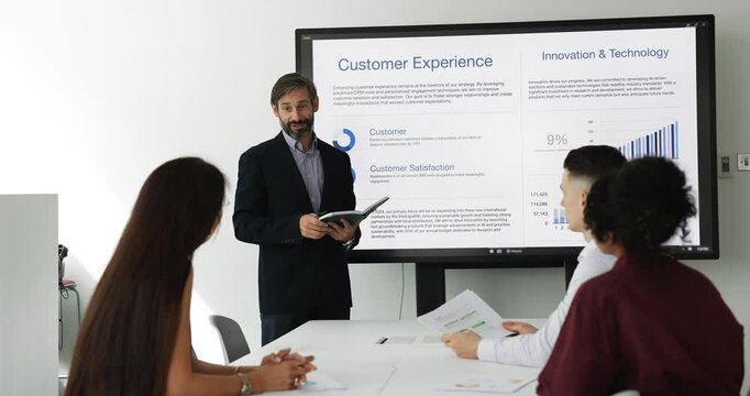 Middle-aged Portuguese team leader gives presentation, sharing key business insights, standing in front of seminar, training or negotiations participants, explaining slides details displayed on screen