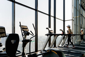 Woman walking on treadmill in high-rise fitness gym with panoramic window view, morning workout...