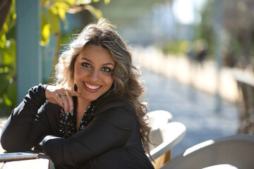 Portrait of young, pretty, blonde woman, wearing a black dress with sequins, posing looking at camera, smiling and happy, while sitting on a stool, in an outdoor park.