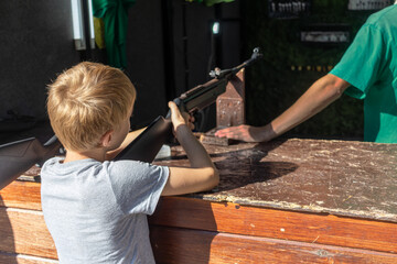 Young boy aiming an air rifle at carnival shooting range, concentrating on target. setting is typical fairground with wooden counter and visible attendant