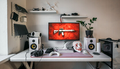 Gaming setup with monitor displaying rifle, speaker system, headphones, game controller, indoor plant, white desk